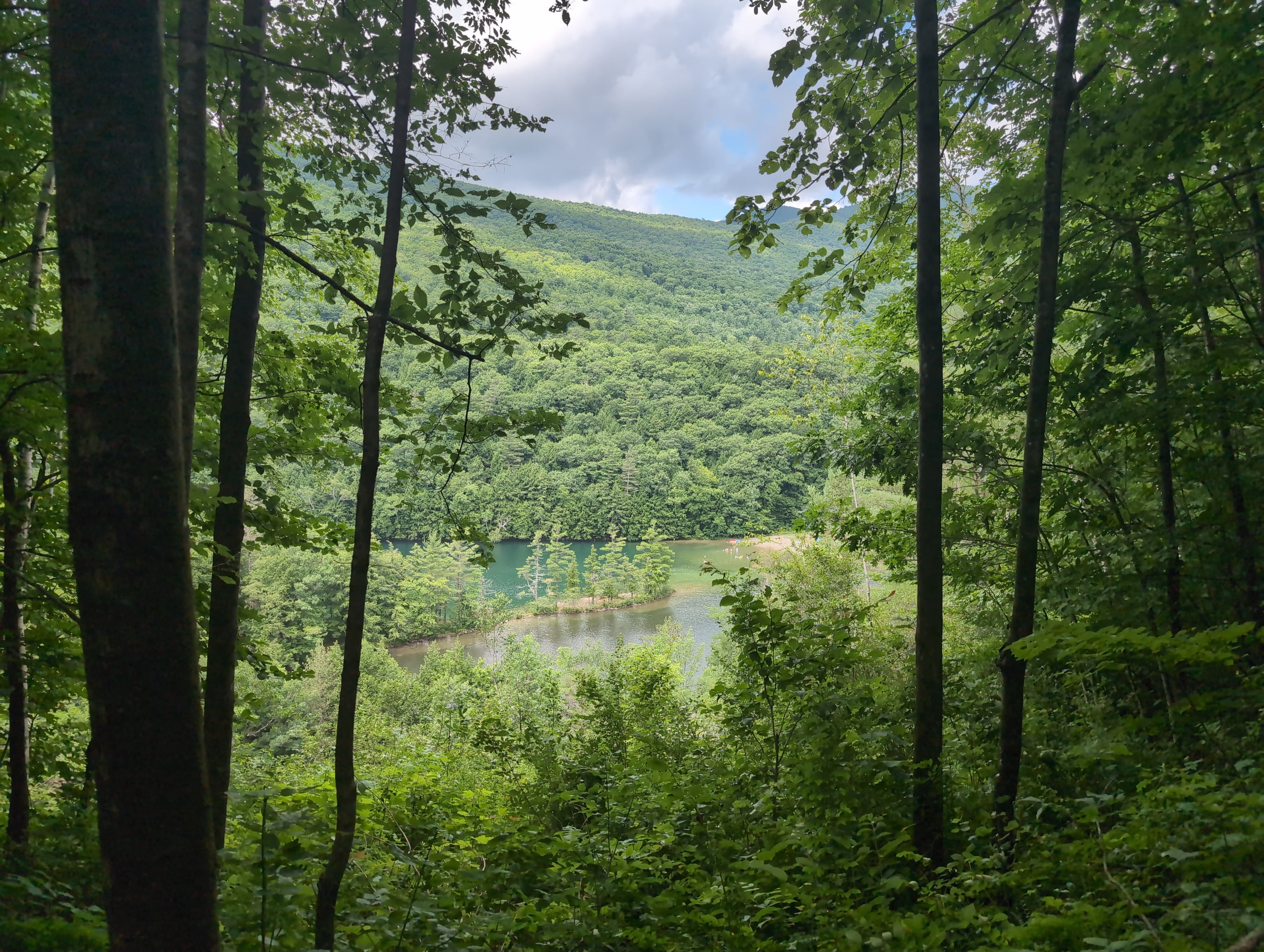 A photo of a lake, taken from a high adjacent hill. Trees frame the image, and an island stretches horizontally through the lake. The water above the island in the image is a deep green color, while that below it is brown. Snatches of blue sky are visible between the clouds over the slope of the next hill beyond the lake.